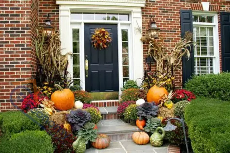 A welcoming fall porch decorated with large orange pumpkins, various gourds, vibrant mums in red and yellow, tall cornstalks, and a festive autumn wreath on a dark blue front door of a brick house.
