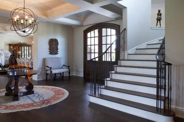 An image demonstrating "Foyer Decorating Ideas," highlighting a sophisticated entryway with an ornate chandelier, a large round table, and a sweeping staircase with dark treads and a black metal banister.
