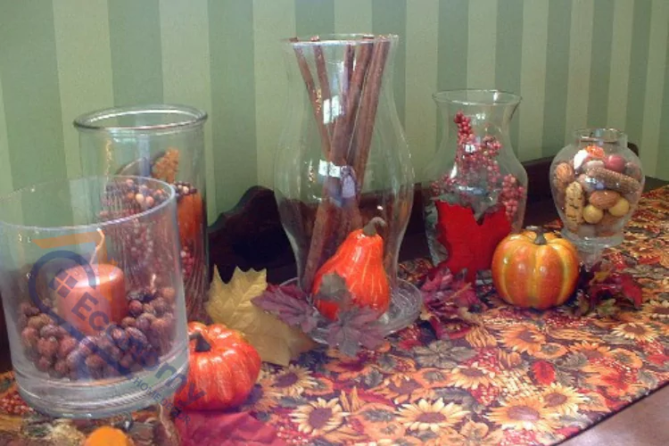 A close-up of a table decorated for autumn, featuring several glass hurricanes and vases filled with seasonal items like cinnamon sticks, berries, nuts, and faux pumpkins, all resting on a patterned fall-themed table runner.