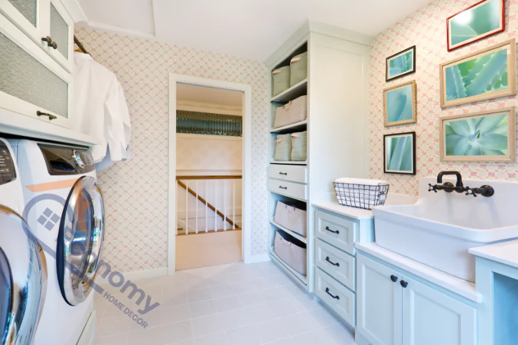 Laundry room decor with soft pink patterned wallpaper, light blue cabinetry, a large utility sink, and wall art with botanical prints, alongside a washing machine and dryer.