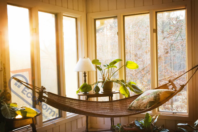 A cozy sunroom bathed in warm sunlight, featuring a woven hammock with a patterned pillow, a small side table, and multiple green houseplants.