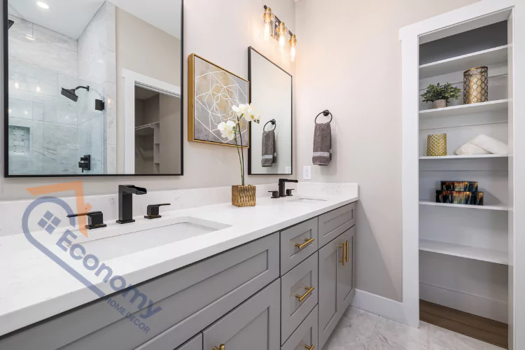 A modern bathroom with a double vanity and a built-in shelf for storage, featuring gray cabinets, gold hardware, and a large shower in the background.