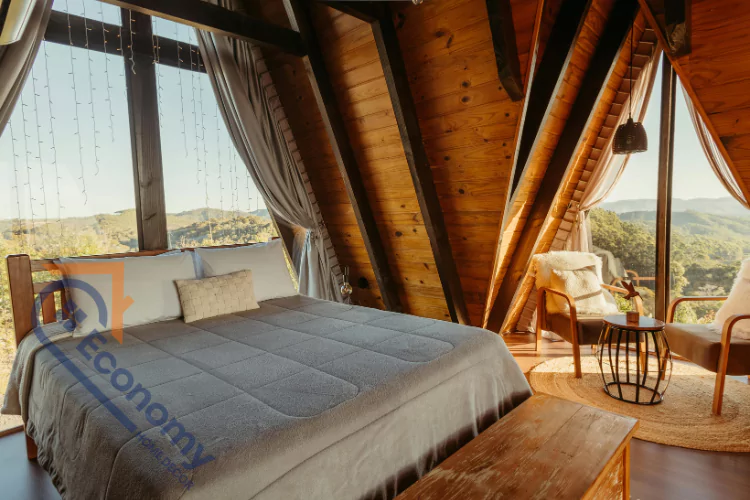 A beautiful cottage bedroom with a high, slanted wooden ceiling and large windows offering a panoramic view of mountains, featuring a bed with a gray duvet and white pillows.