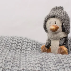 Cozy winter decor: Close-up of a small penguin figurine wearing a grey knitted hat, nestled in a thick grey knit blanket.
