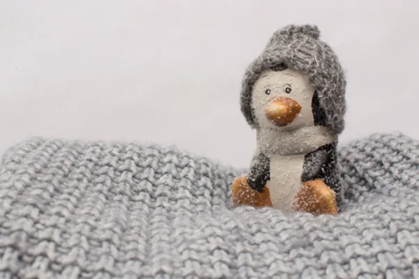 Cozy winter decor: Close-up of a small penguin figurine wearing a grey knitted hat, nestled in a thick grey knit blanket.