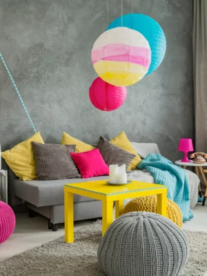 Vibrant and eclectic living room with a grey textured wall, colorful paper lanterns, yellow coffee table, and pink and grey poufs.