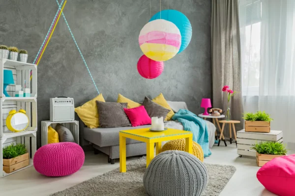 Vibrant and eclectic living room with a grey textured wall, colorful paper lanterns, yellow coffee table, and pink and grey poufs.