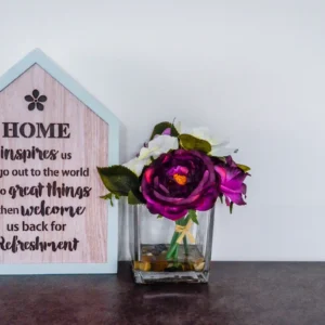 A wooden house-shaped plaque with an inspirational quote next to a square glass vase with purple and white flowers.