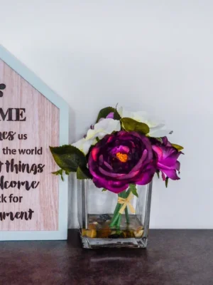 Home Inspires Us A wooden house-shaped plaque with an inspirational quote next to a square glass vase with purple and white flowers.