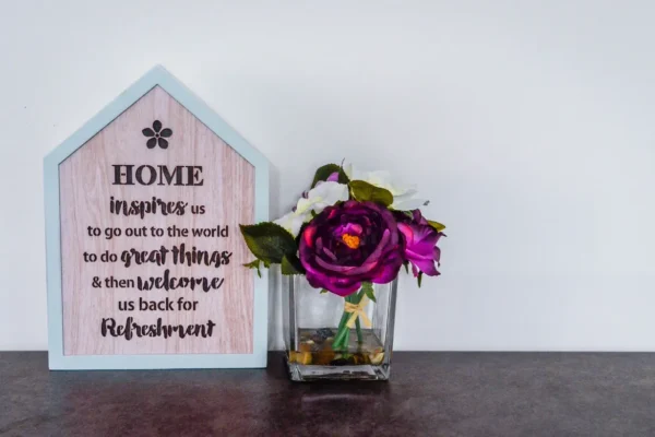 A wooden house-shaped plaque with an inspirational quote next to a square glass vase with purple and white flowers.