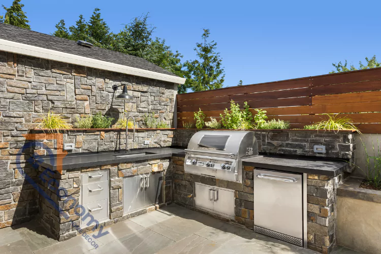 A modern outdoor kitchen with a stone facade, featuring a built-in stainless steel grill, sink, and refrigerator, with a stone wall and wood fence in the background.