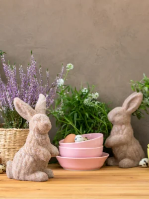 Spring and Easter table decor with two flocked bunny figurines, fresh eggs, and potted plants on a wooden surface.