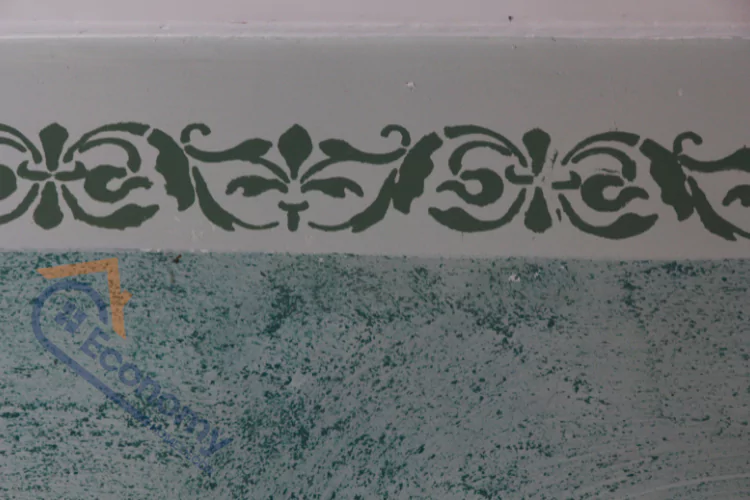 A close-up of a wall with a two-tone paint job, featuring a decorative stencil border with a damask-like pattern in dark green at the top.