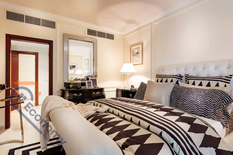 A sophisticated bedroom with a black and white patterned comforter on a bed with a large tufted headboard, and a mirrored vanity area to the left.