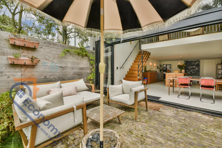 A cozy urban outdoor living space with wood-framed seating, light cushions, a low wooden table, and a striped umbrella over a brick patio.