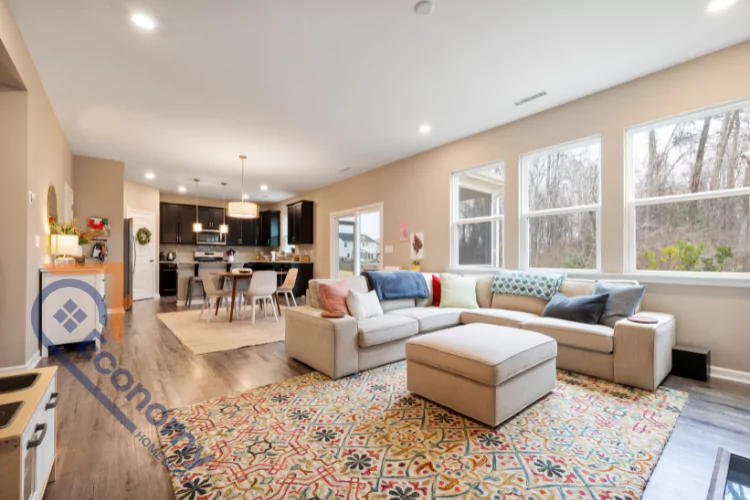 A spacious open-concept living room and kitchen in a single wide mobile home, with light-colored walls and a sectional sofa on a vibrant patterned rug.