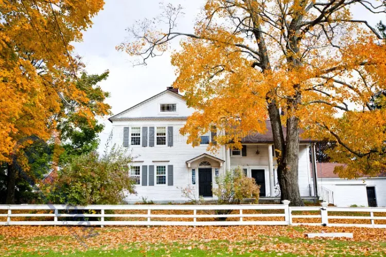 An aerial photograph of a large white house on a green lawn, surrounded by mature trees with bright orange and yellow fall leaves, with a white picket fence in the foreground.