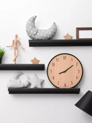 Three black floating shelves decorated with a large wooden clock, plush moon and star pillows, a mannequin, and a small picture frame.