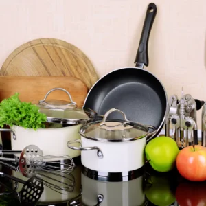 Complete kitchen set featuring white saucepans with glass lids, a large black frying pan, wooden cutting boards, and assorted stainless steel utensils.