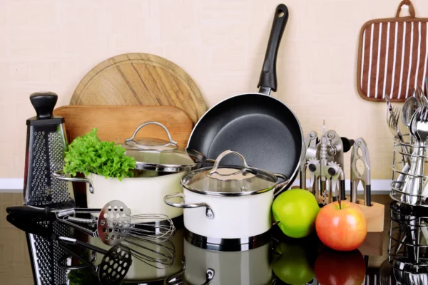 Complete kitchen set featuring white saucepans with glass lids, a large black frying pan, wooden cutting boards, and assorted stainless steel utensils.