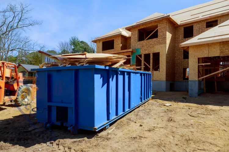 A wide, blue roll-off dumpster filled with construction debris, lumber, and plywood sits on the dirt ground in front of a partially completed wood-framed multi-unit housing renovation project.