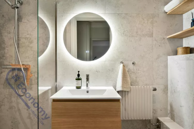 A contemporary bathroom with a round, backlit LED decorative mirror hanging over a light wood vanity.