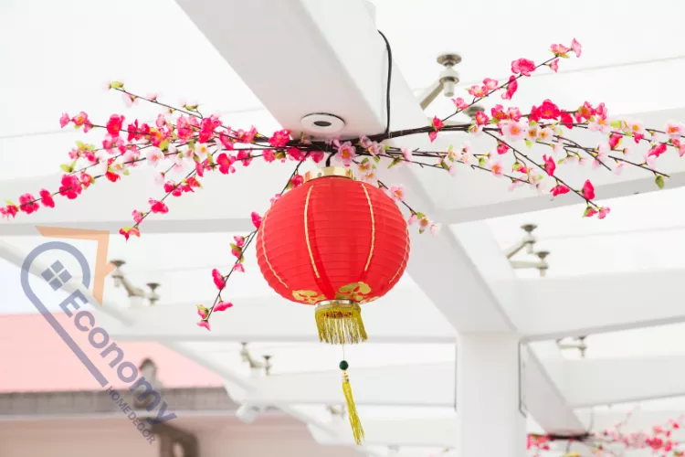 A vibrant red paper or silk lantern with a gold tassel hanging from a ceiling beam, surrounded by pink artificial cherry blossoms.