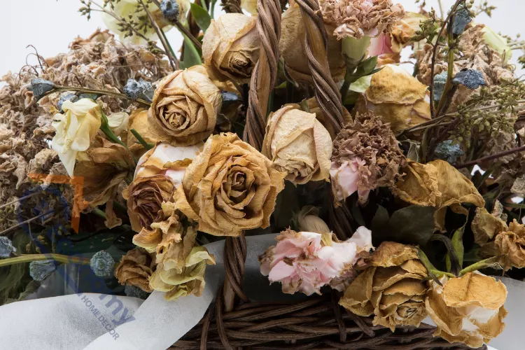 A close-up of a wicker basket overflowing with a large, rustic arrangement of faded tan and yellowish dried rose blooms, along with various other light-colored preserved flowers and foliage.