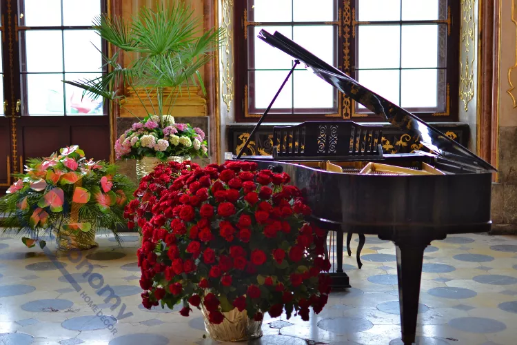 A beautiful black grand piano in a formal setting, surrounded by massive arrangements of red and pink flowers.