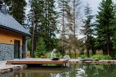 Backyard oasis with a small wooden deck next to a tranquil pond, featuring a modern rustic house and a peaceful, wooded natural setting.
