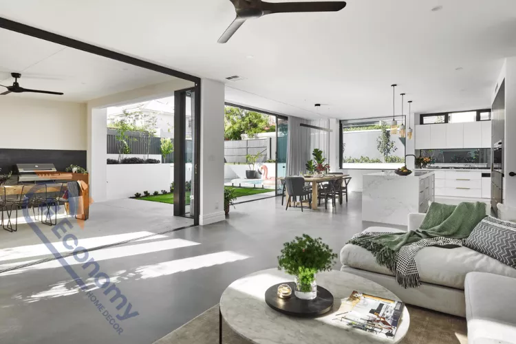Bright, modern, open-plan living space with light gray polished concrete floors extending through the living, dining, and kitchen areas, opening to an outdoor patio and pool area through large folding glass doors.