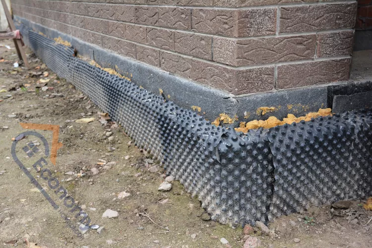 A worker performing a chemical injection for damp proofing on an exterior brick wall, wearing work gloves and using specialized equipment to treat a row of holes above the ground line.