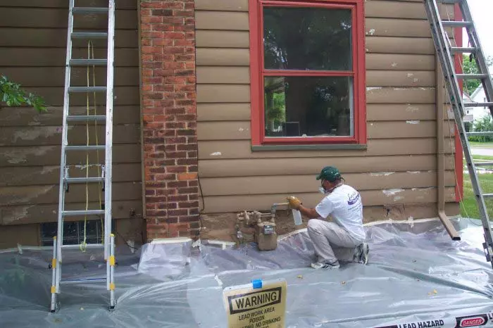 A professional painter from Lang's Painting applying a white coat of paint to the wooden trim of a large, dark-colored residential house exterior.