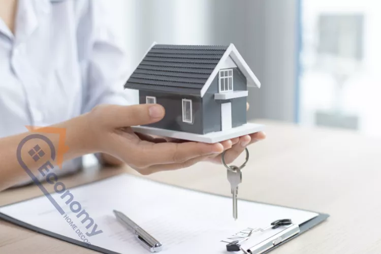 A person's hands holding a miniature model of a gray house with a dark roof over a clipboard with a pen, key, and documents, symbolizing home financing or Infonavit points.