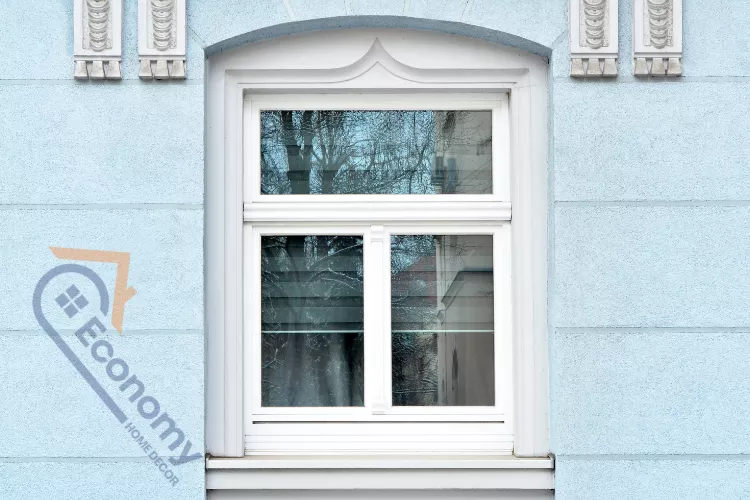 A close-up of a classic white-framed exterior window featuring ornate decorative molding and an arched top detail, set against a light blue textured wall with horizontal rustication lines.