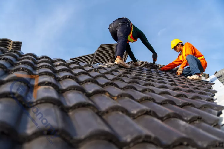Low-angle view of two construction workers installing dark, curved roof tiles on a house under a blue sky.